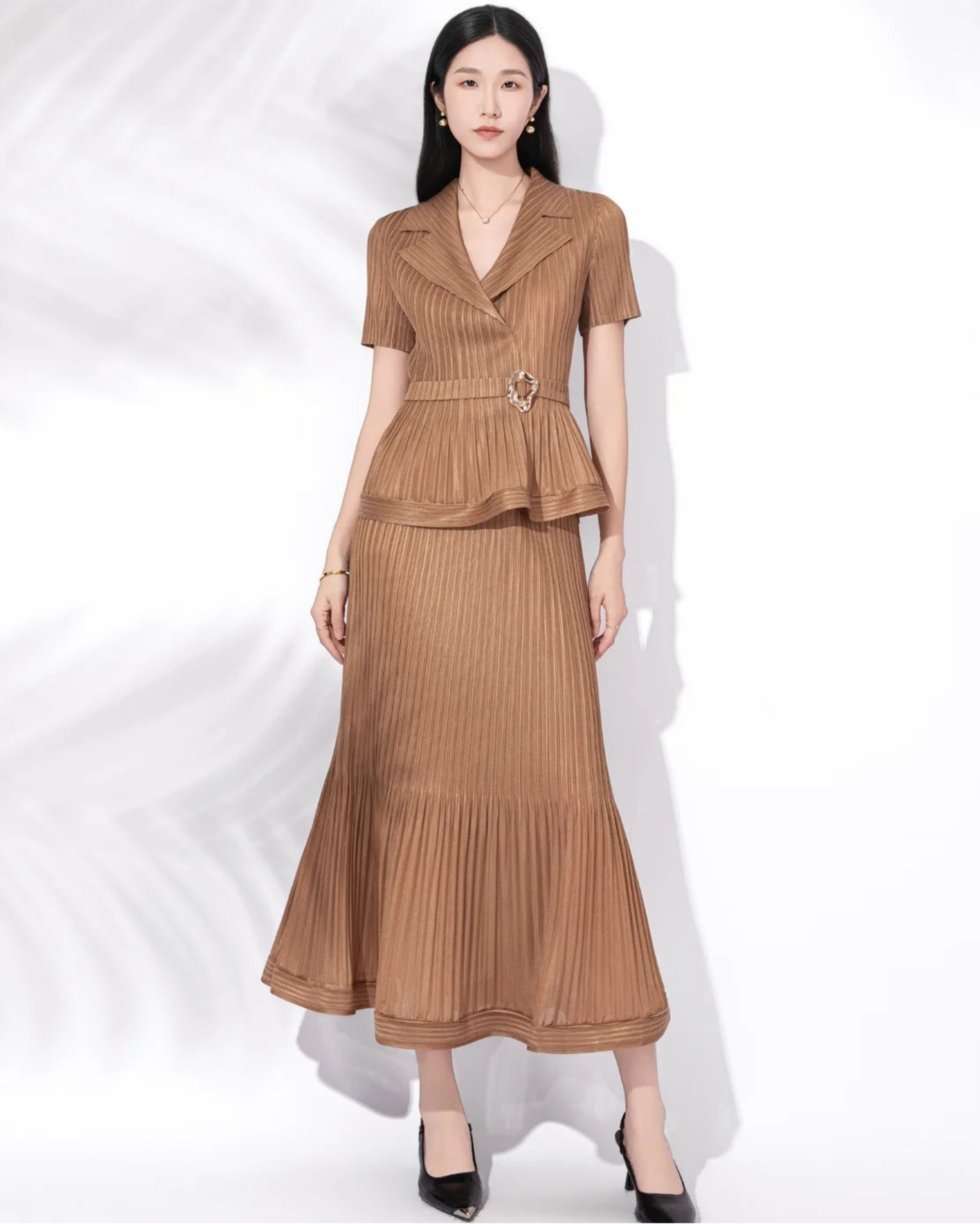 A woman modeling a two-piece brown pleated set consisting of a short-sleeve collared top with a peplum waist and a matching pleated maxi skirt, styled with black heels against a white background with soft leaf shadows.