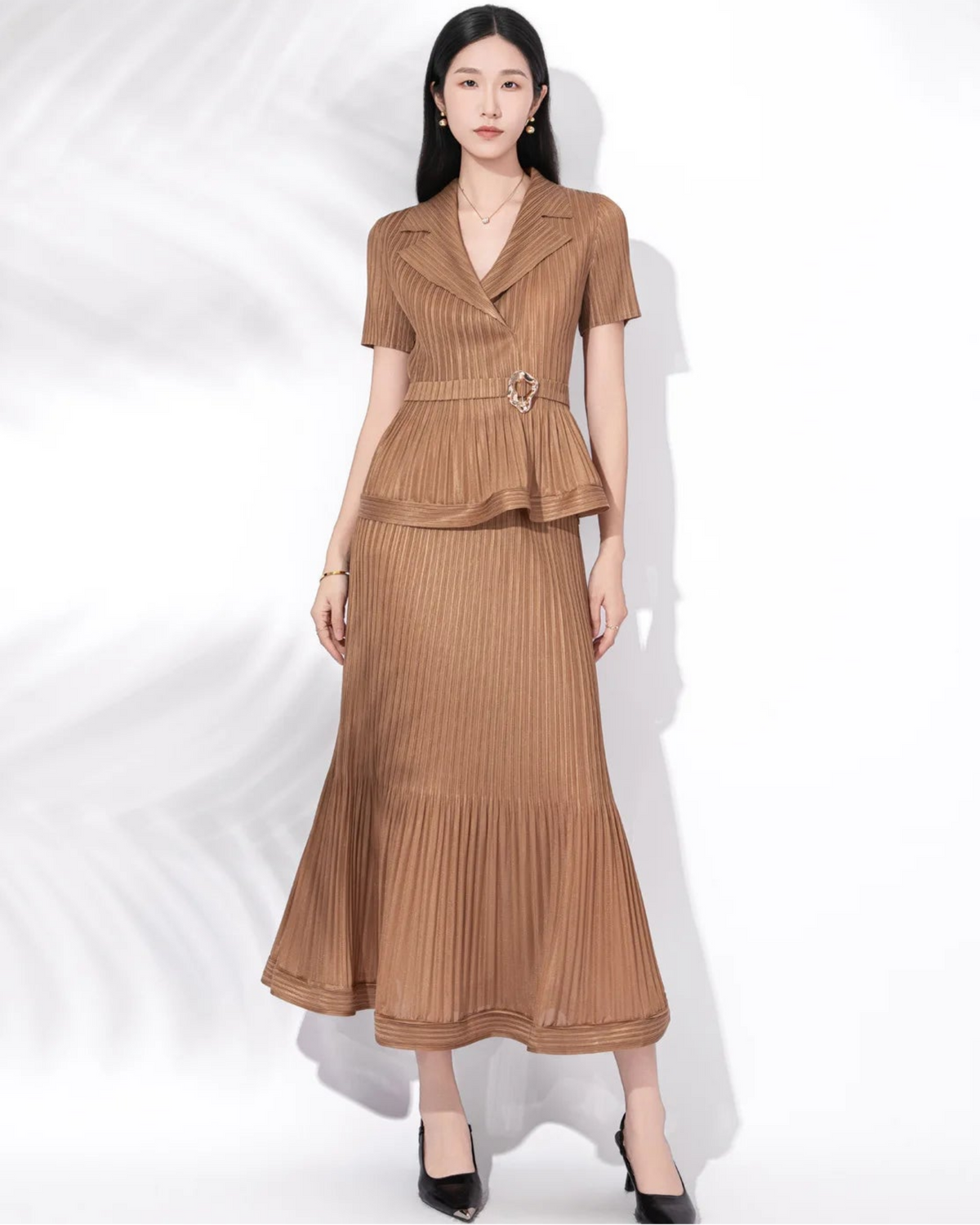 A woman modeling a two-piece brown pleated set consisting of a short-sleeve collared top with a peplum waist and a matching pleated maxi skirt, styled with black heels against a white background with soft leaf shadows.