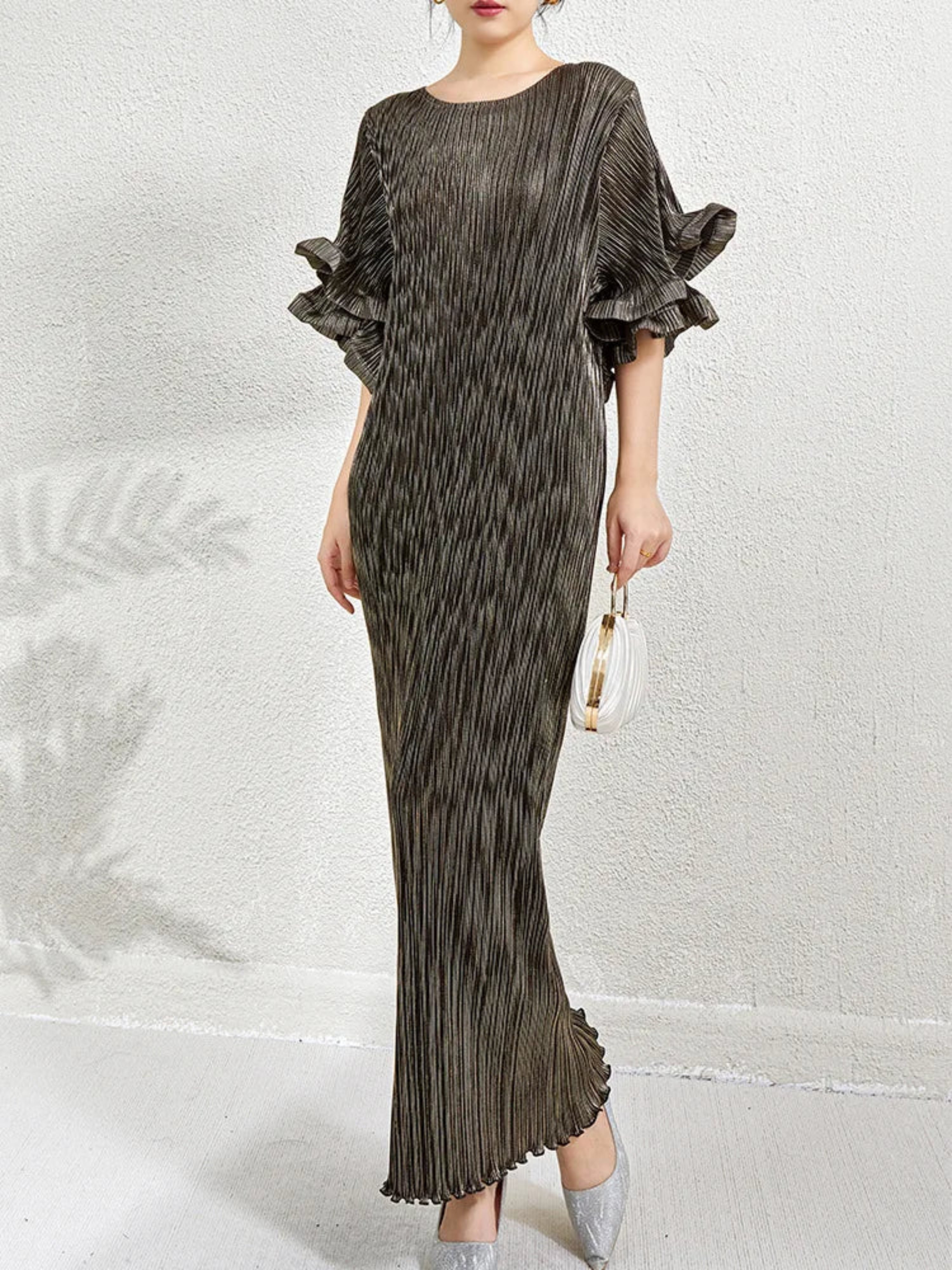 A woman models a floor-length pleated dress with dramatic ruffled elbow-length sleeves in a dark taupe-brown color, holding a small white handbag against a textured white wall.