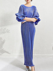 A woman models a royal blue pleated maxi dress with voluminous ruffled elbow-length sleeves, set against a textured off-white wall.