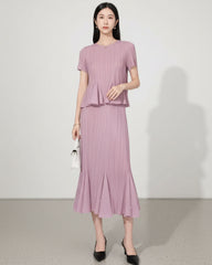 A woman models a matching two-piece pleated set in a dusty lavender-pink color, featuring a short-sleeve top with a ruffled hem and a high-waisted pleated skirt with small pearl-like embellishments, paired with black heels.