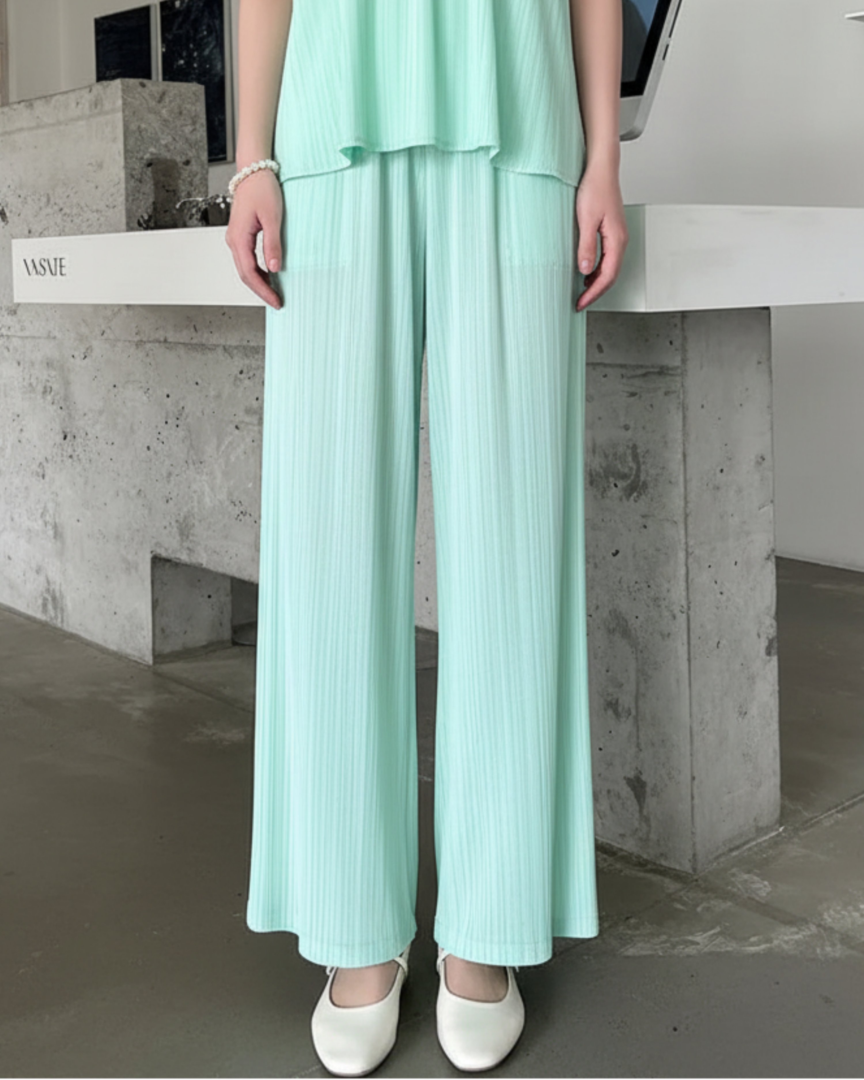 A person wearing a mint green two-piece matching set with a sleeveless top and wide-leg pleated pants, paired with white Mary Jane flats, standing in a modern interior with a concrete counter and a laptop in the background.