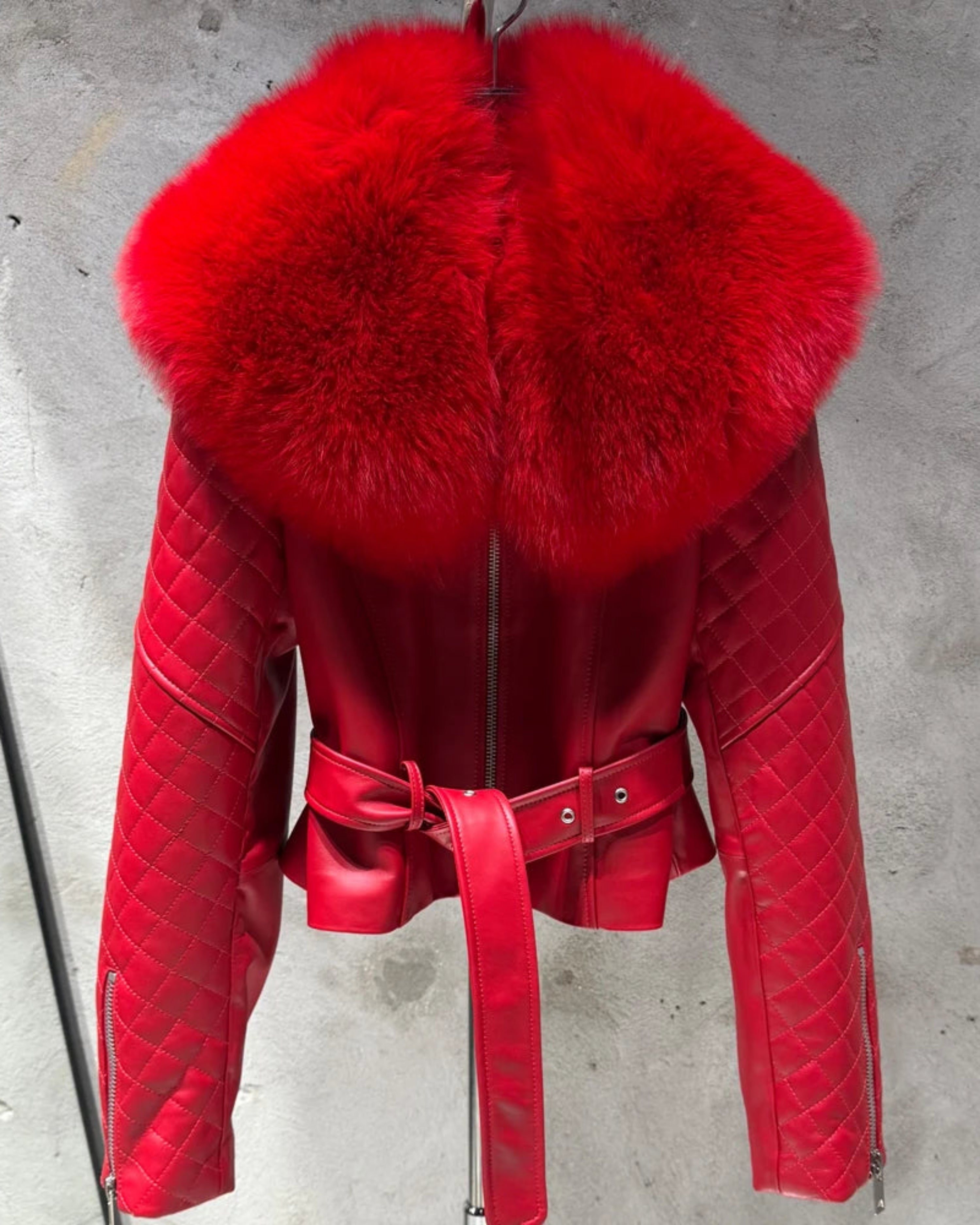 A bright red quilted leather jacket with a large fluffy red fur collar, a matching red belt, and silver zippers, shown hanging against a gray concrete wall.